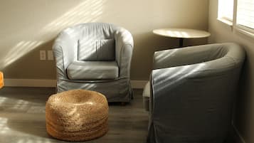 Una habitación con un sillón gris, un taburete redondo tejido, una mesita redonda y una ventana con persianas.