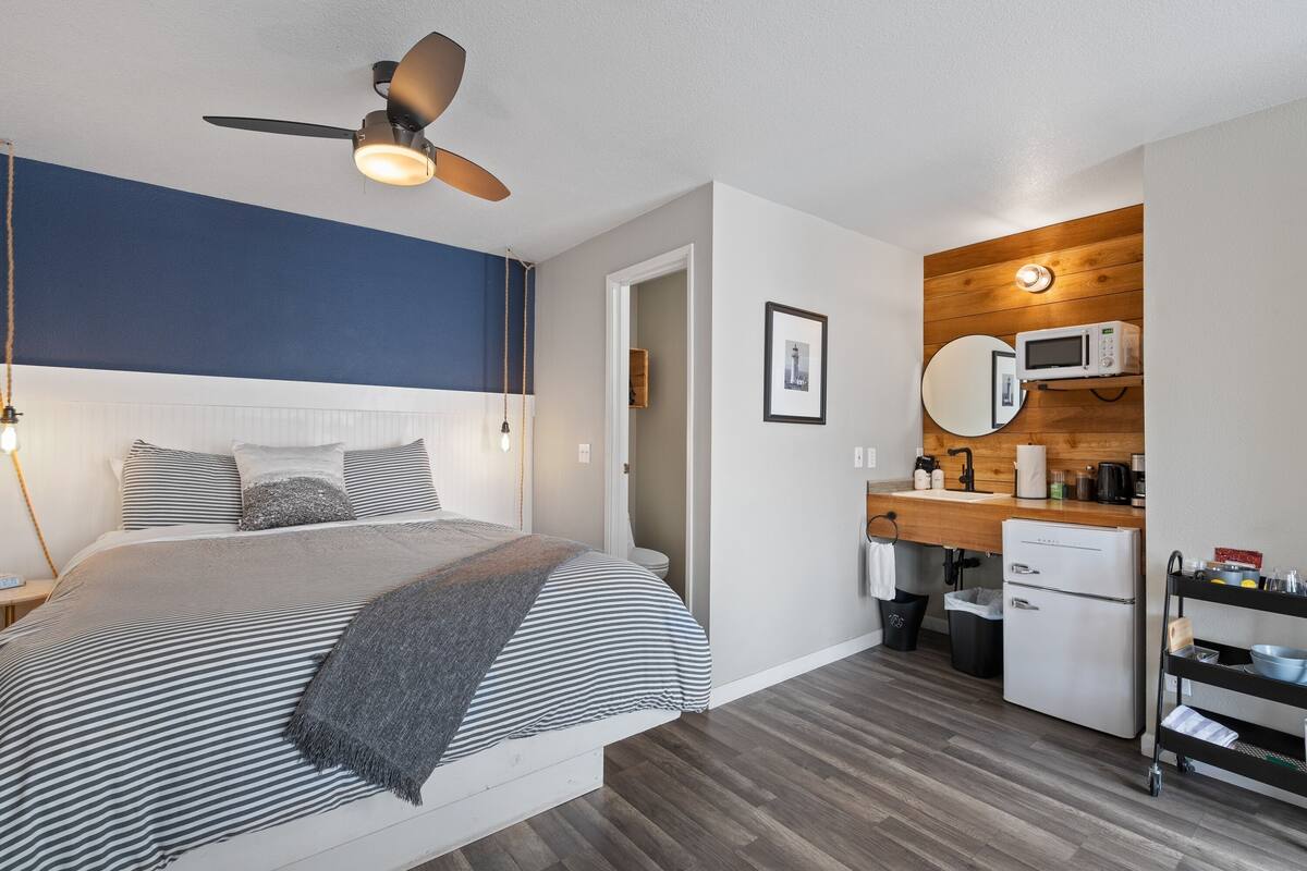 A bedroom with a bed, bedside table, and a ceiling fan. Adjacent to the bedroom is a small kitchenette with a microwave, sink, and refrigerator.