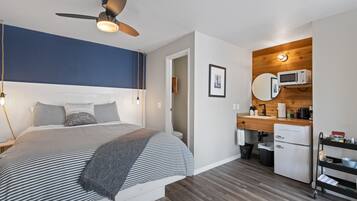 Une chambre comprenant un lit, une table de chevet et un ventilateur de plafond. À côté de la chambre se trouve une petite kitchenette équipée d’un four à micro-ondes, d’un évier et d’un réfrigérateur.