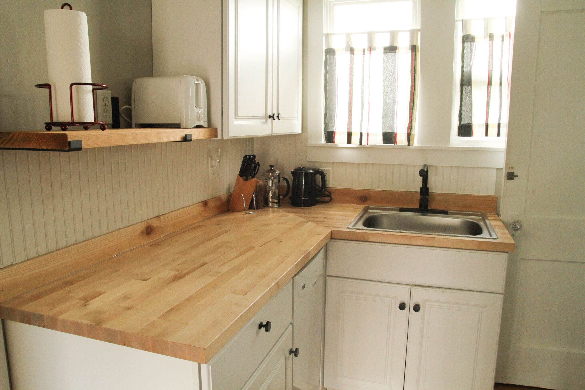 Une cuisine avec des plans de travail en bois, des meubles blancs, un évier et une fenêtre avec des rideaux.