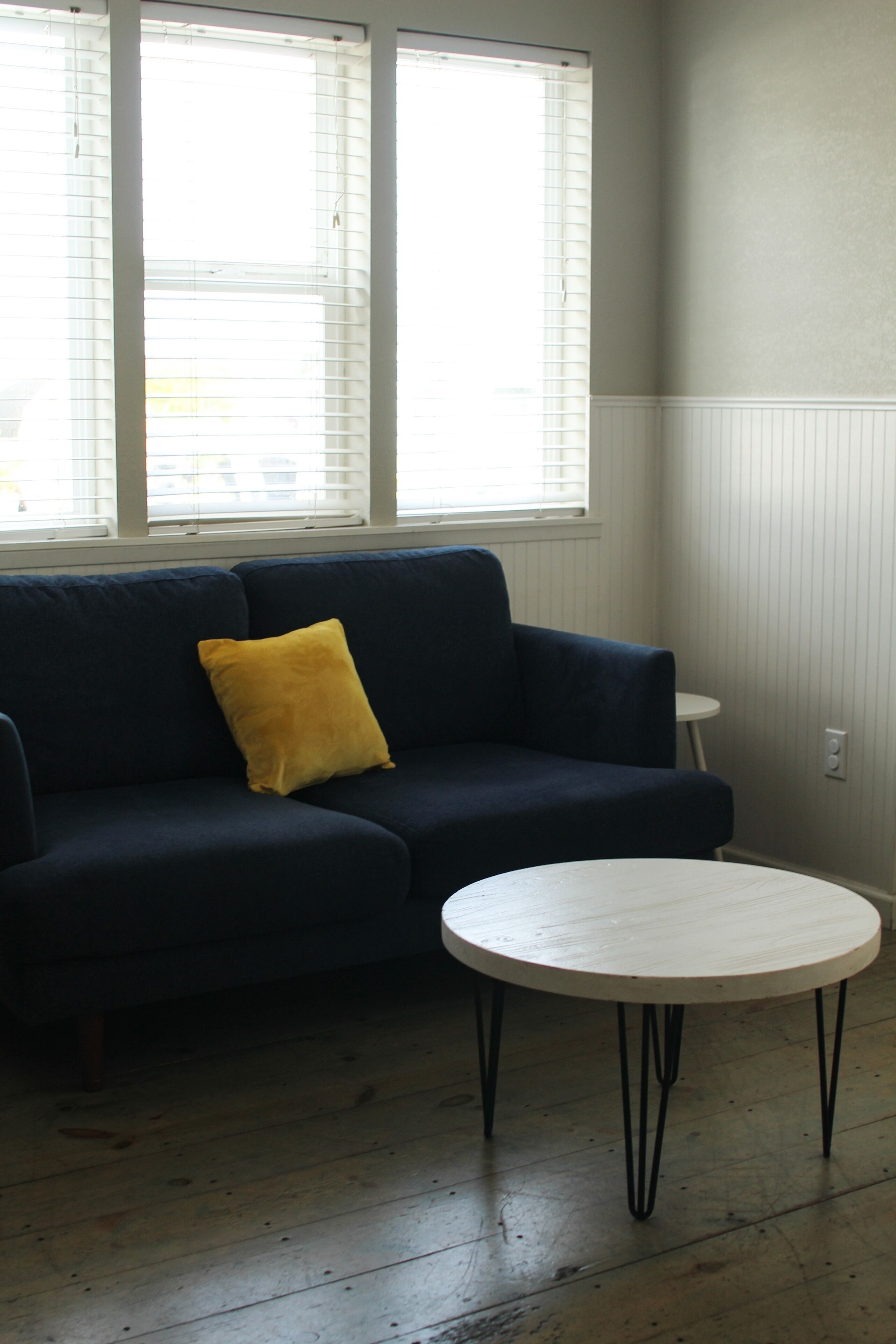 Une pièce avec un canapé bleu foncé, un coussin jaune et une petite table ronde blanche aux pieds noirs.