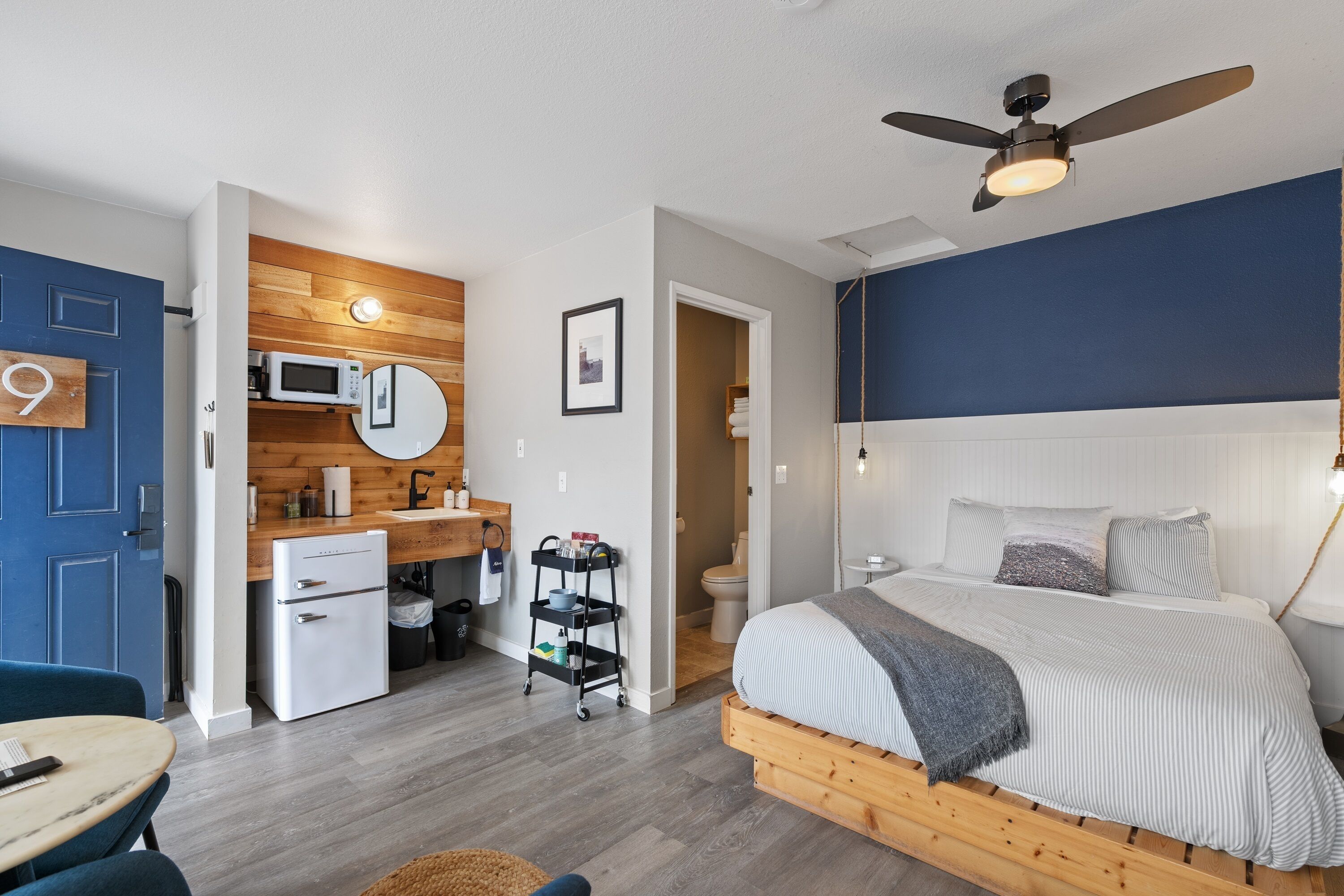 A compact hotel room with a bed, a small kitchenette, a microwave, and a ceiling fan.