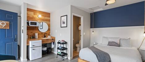 A compact hotel room with a bed, a small kitchenette, a microwave, and a ceiling fan.