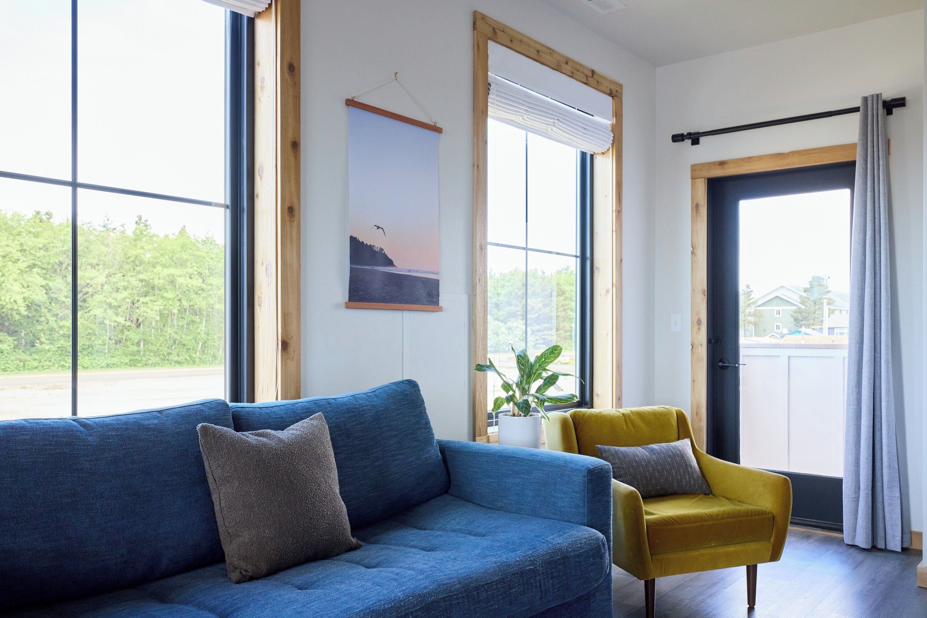 Uma sala de estar moderna com um sofá azul, uma poltrona amarela e uma grande janela com vista para o verde.