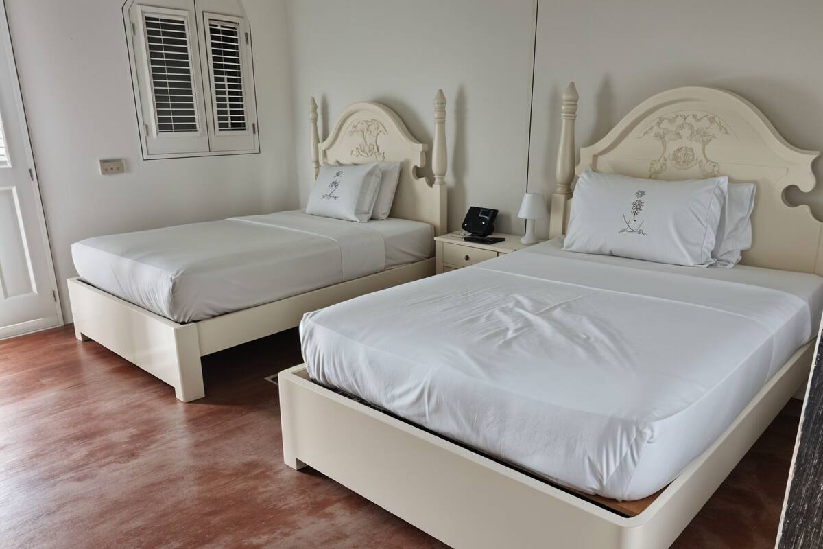 A bedroom with two white beds, a nightstand with a lamp, and a window with shutters.