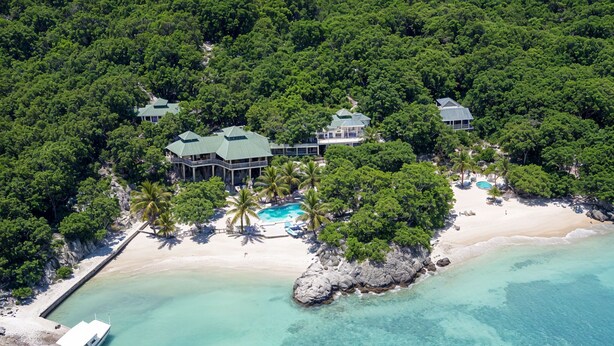 Vista aérea de un resort tropical con varias edificaciones, una playa y un mar turquesa y transparente.