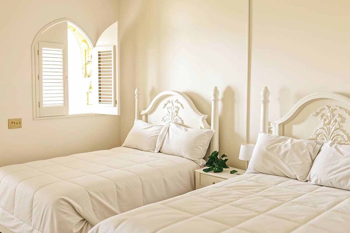 A bedroom with two beds, wooden beams on the ceiling, a window with shutters, and a green rug.