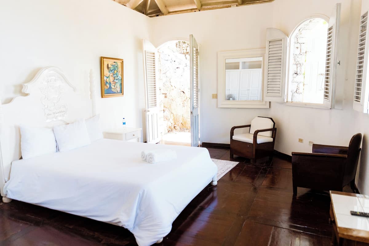 A bedroom with a large bed, a desk, and a chair. There is a window with shutters and a painting on the wall.