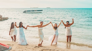 Un grupo de personas con vestidos blancos de pie en una playa, con un barco al fondo.