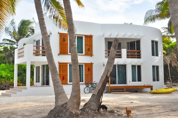 Un edificio blanco de dos pisos con balcones y ventanas de madera, rodeado de palmeras y una playa arenosa.