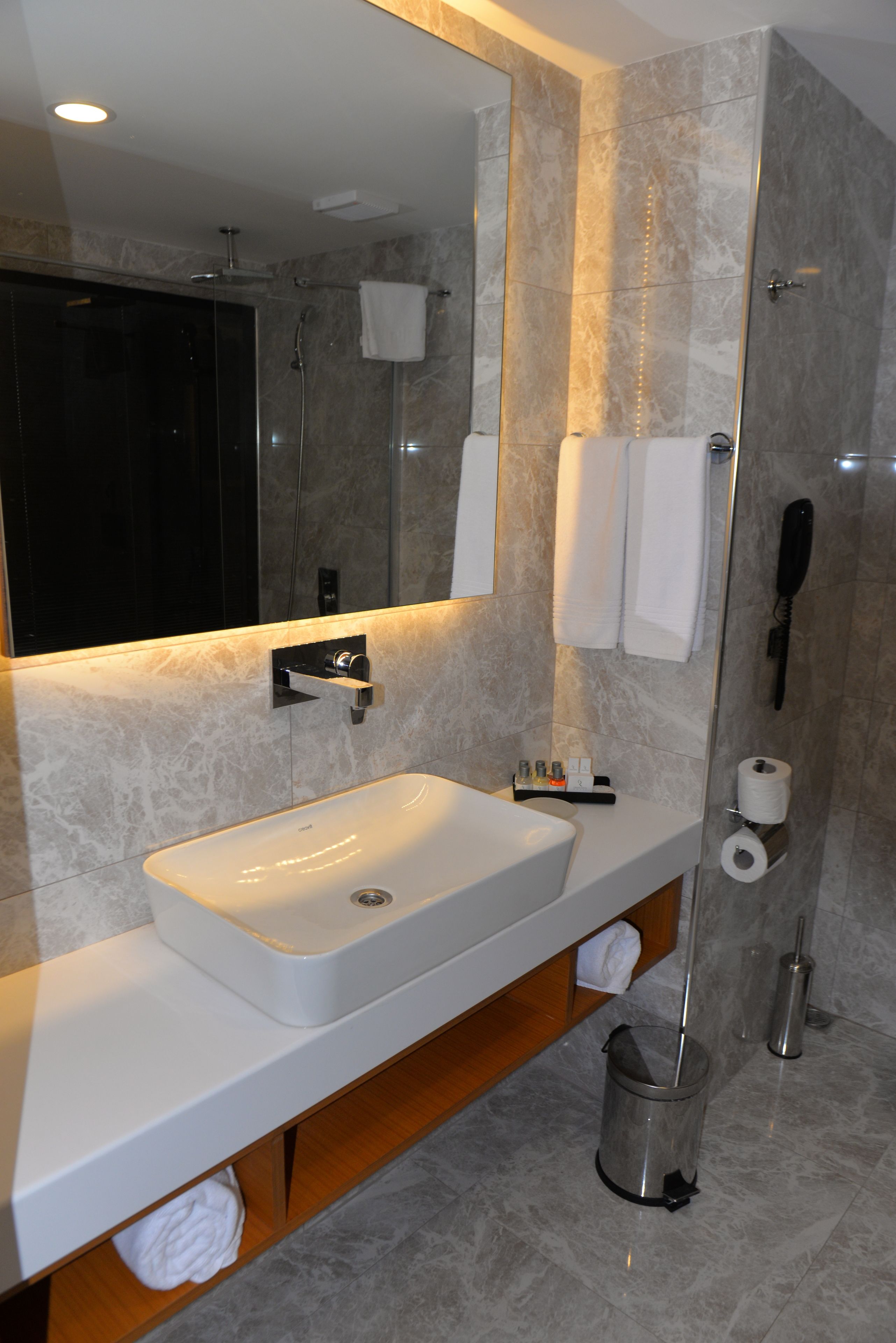 A modern bathroom with a large mirror, a white sink, a shower, and a toilet.