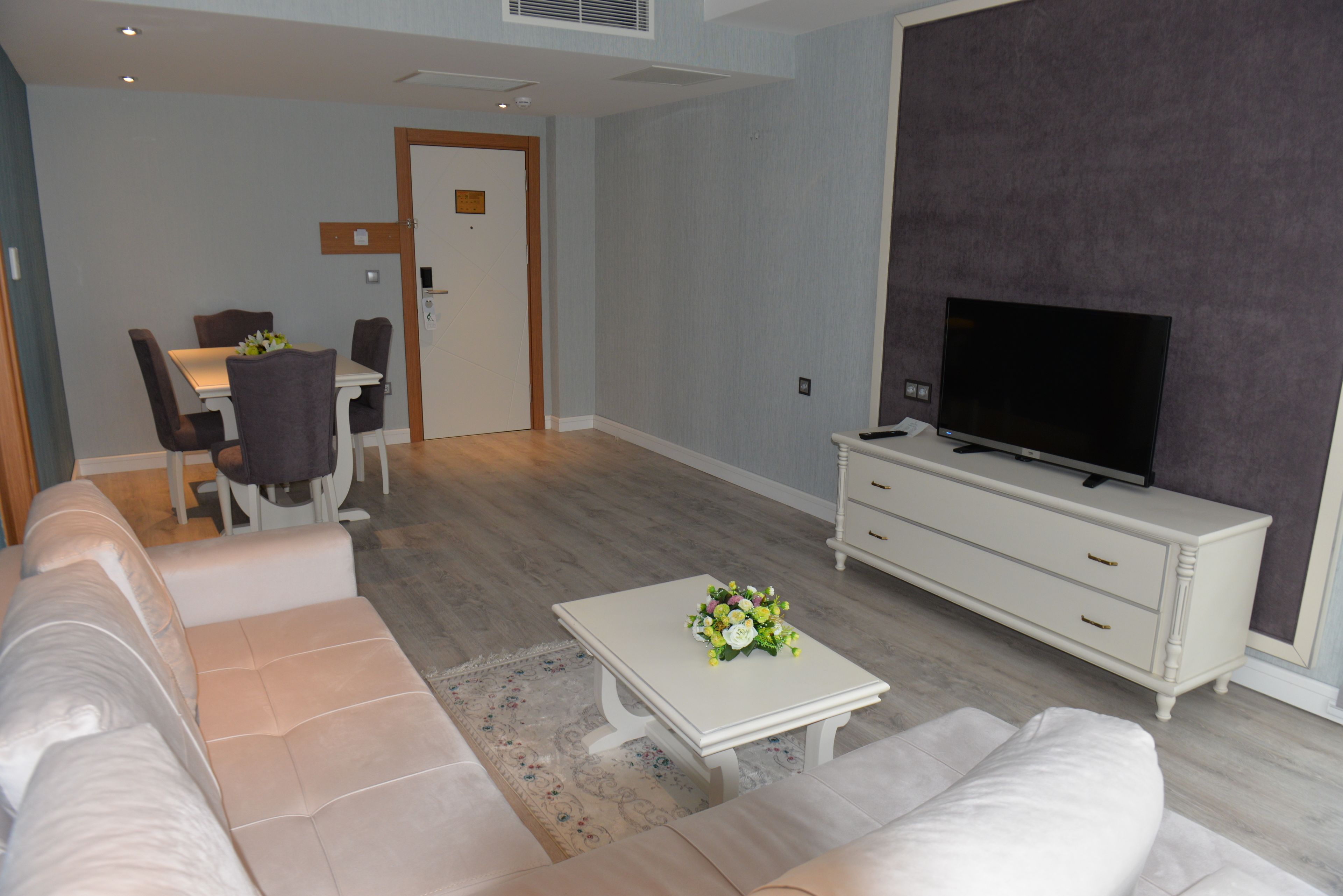 A modern living room with a sofa set, a dining table with chairs, a TV on a dresser, and a door leading to another room.