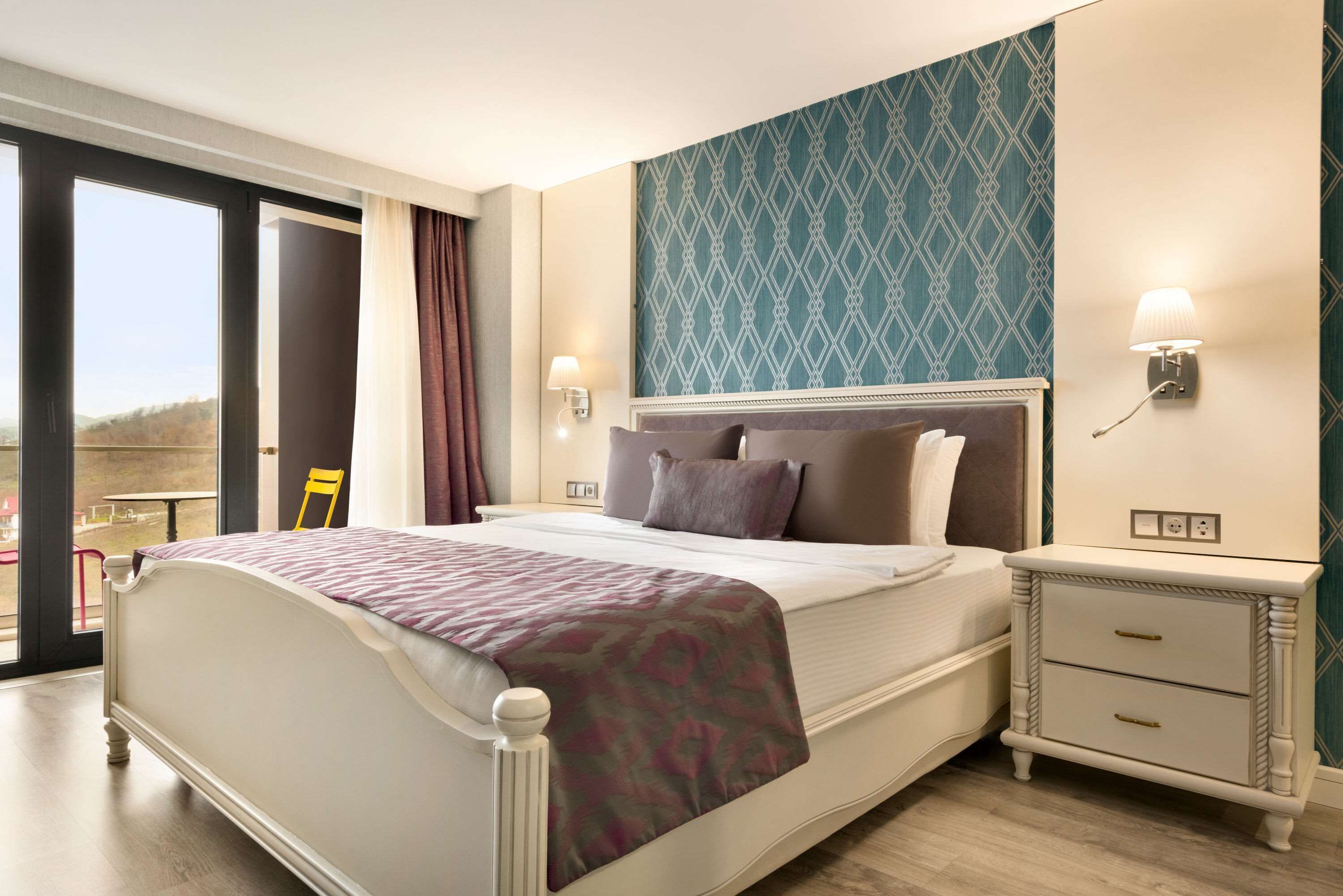 A hotel room with a bed, a nightstand, a window with curtains, and a patterned wall.