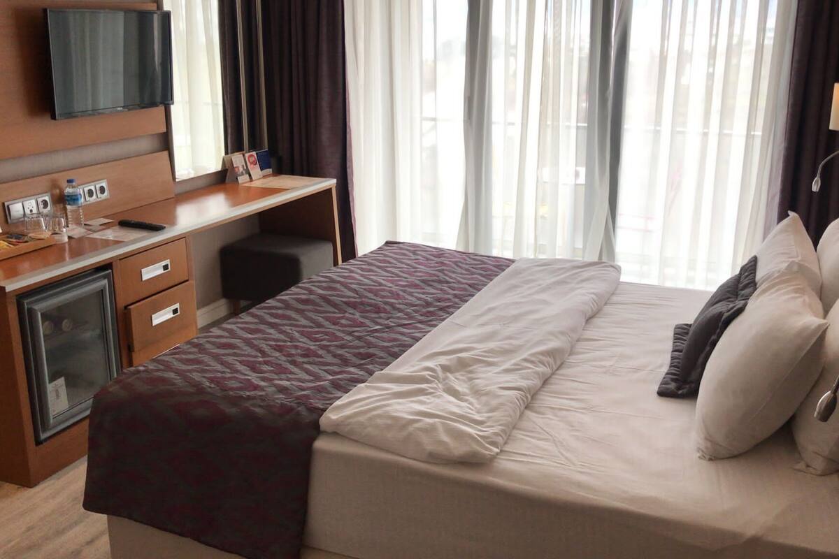 Hotellihuone, jossa on sänky, työpöytä, televisio ja ikkuna, jossa on verhot.