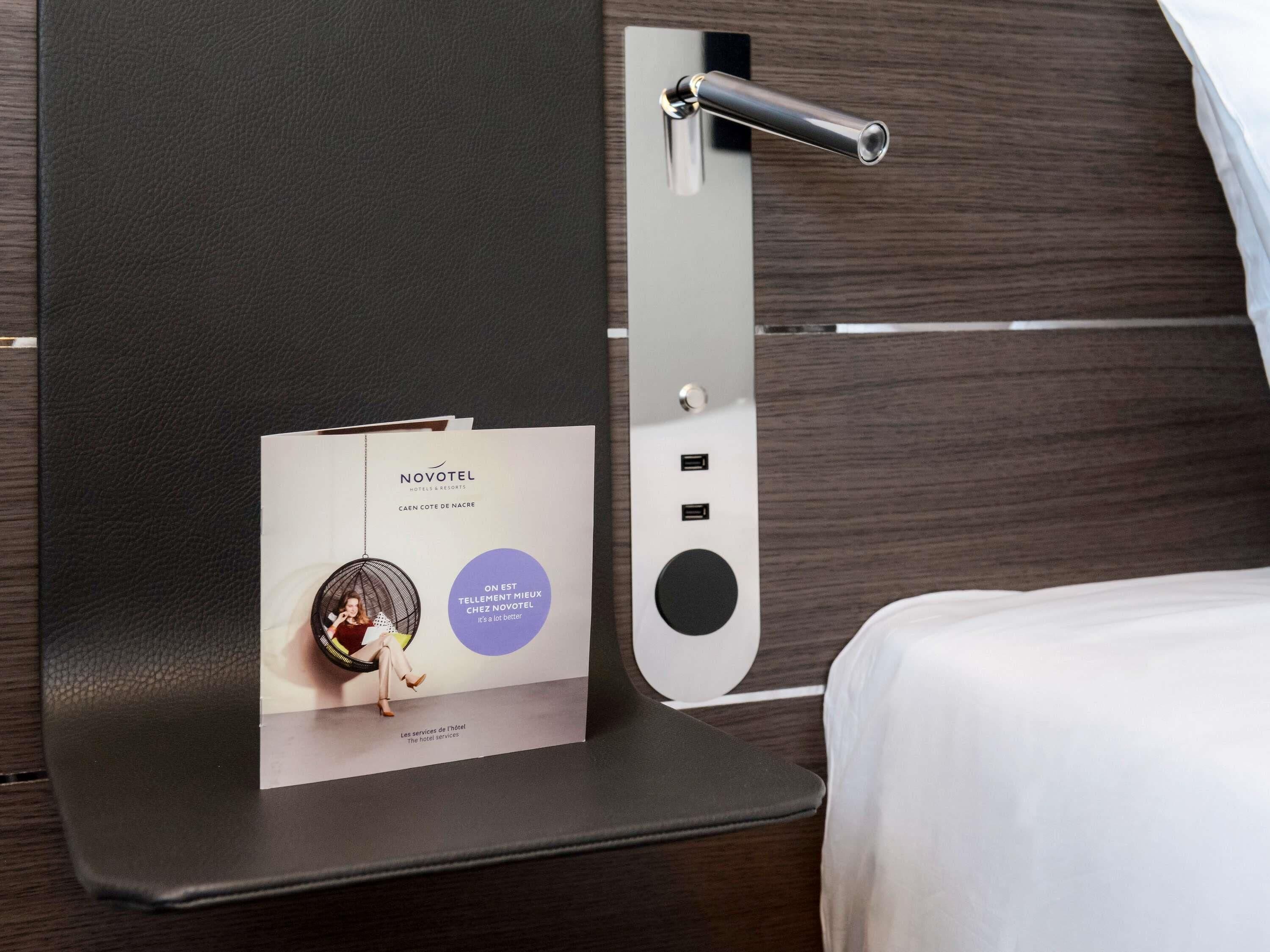 Un folleto del hotel Novotel sobre un atril fijado a la pared, con una cama y un fondo de paneles de madera.