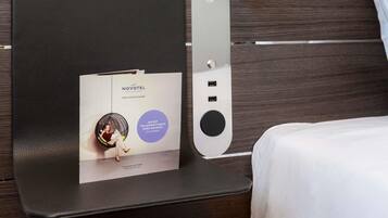 Un folleto del hotel Novotel sobre un atril fijado a la pared, con una cama y un fondo de paneles de madera.