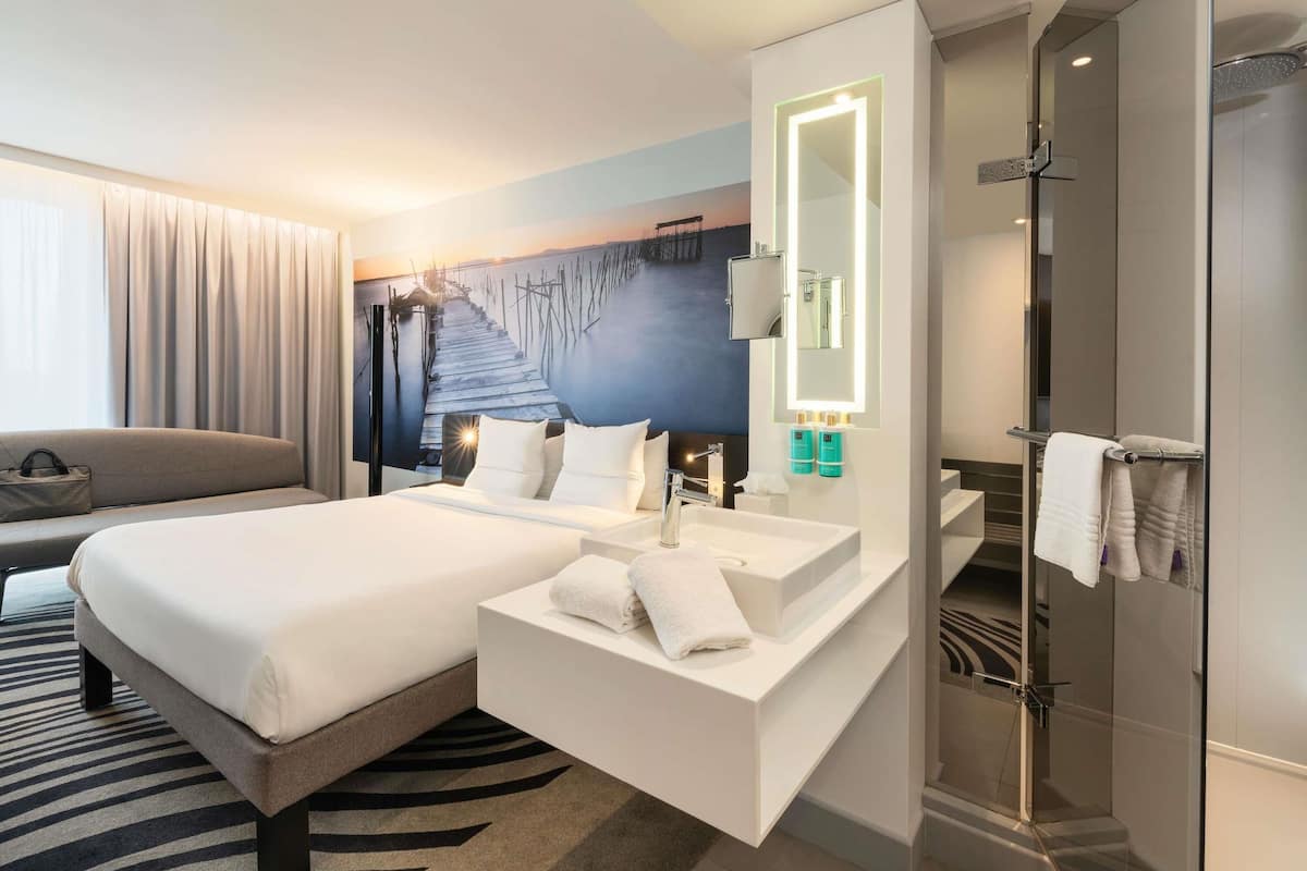 Habitación de hotel moderna con cama, un espejo grande, baño con ducha y vista a un muelle.