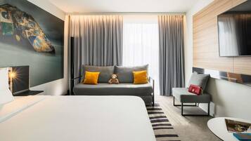 Una habitación de hotel moderna con una cama grande, un sofá con cojines, una mesita y un televisor montado en la pared.