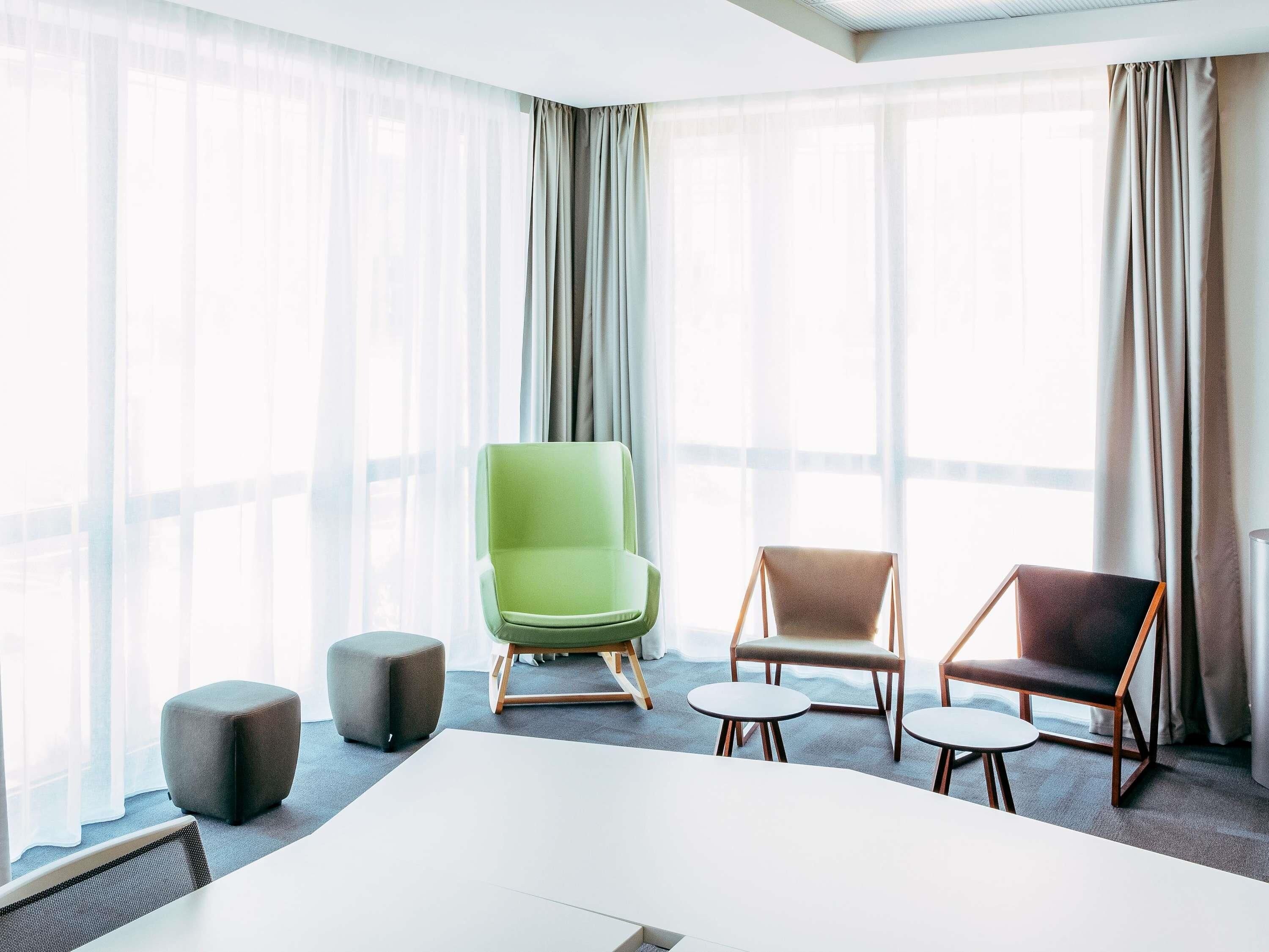 A modern hotel room with a large bed, a green armchair, a brown armchair, a grey ottoman, and a small round table.