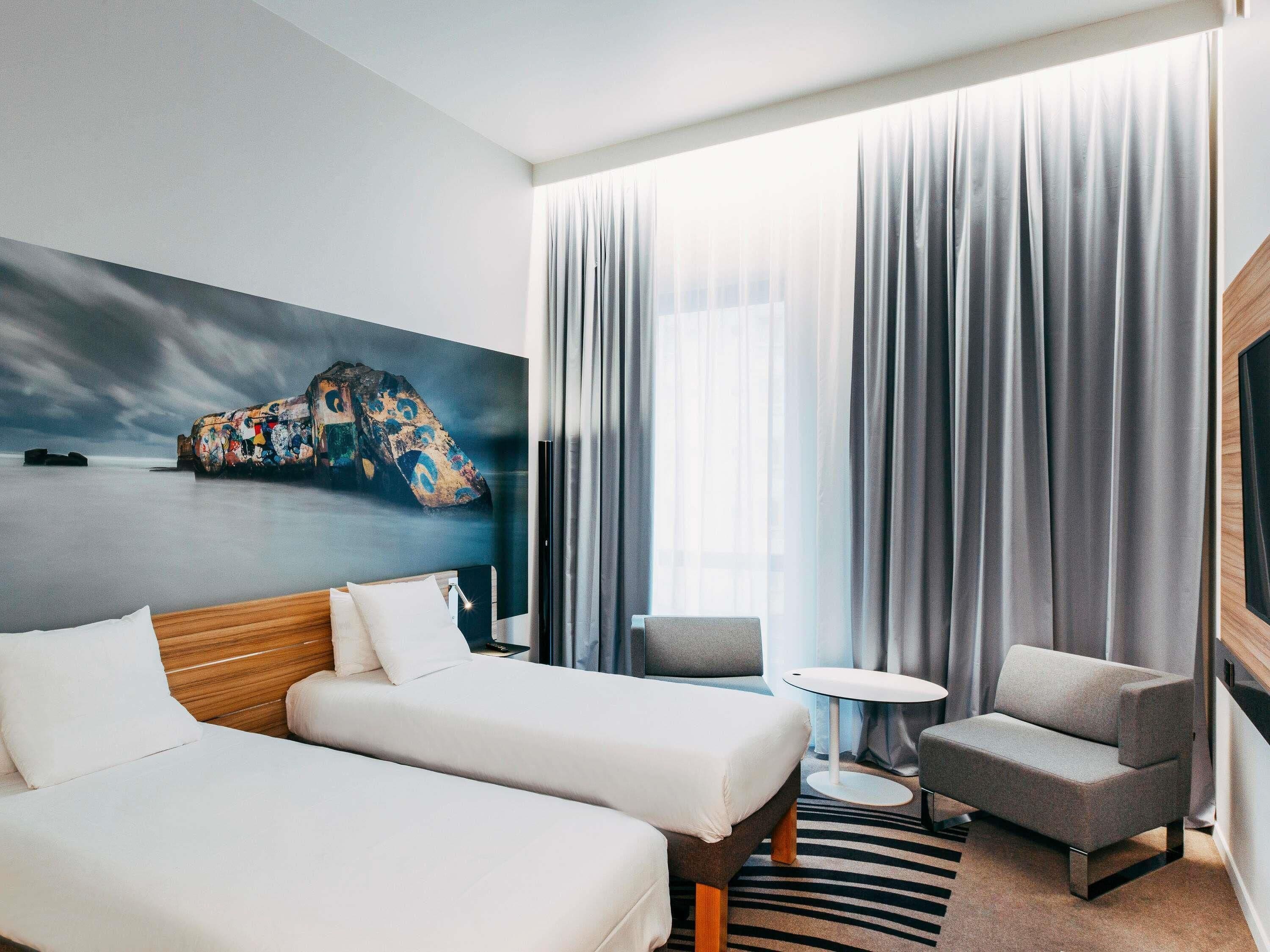 A hotel room with two beds, a small round table, a chair, and a large window with curtains.