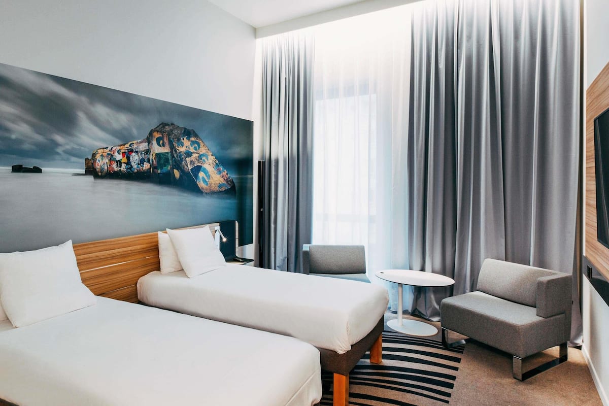Habitación de hotel con dos camas, una mesita redonda pequeña, una silla y un ventanal grande con cortinas.