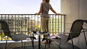 Un balcón con una persona de pie, con vistas a un paisaje urbano. Hay dos sillas y una mesa pequeña con un tazón, una taza y un libro.
