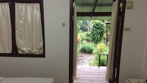 Ein Zimmer mit Bett, einem Vorhangfenster und einer Tür zu einem Außenbereich mit Holzterrasse und viel Grün.