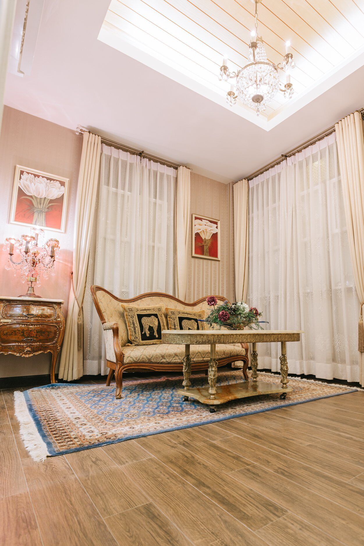 A room with a wooden floor, a sofa, a table with a vase of flowers, and a chandelier.