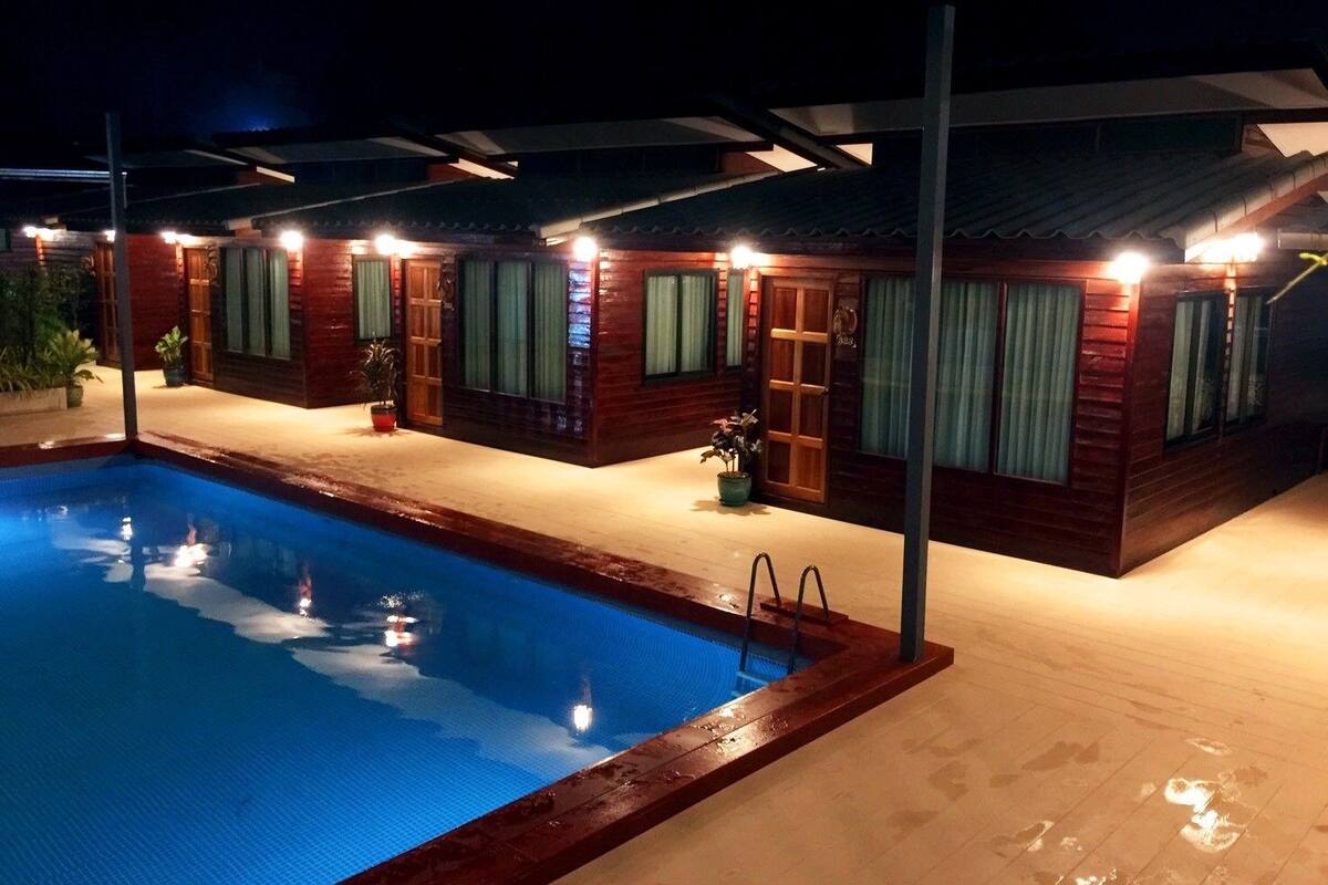 A pool area with wooden cabins, each with a door and windows, and a pool with a light reflecting on the water.