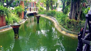Uma via aquática tranquila com fontes decorativas, cercada por vegetação exuberante e uma ponte ao fundo.