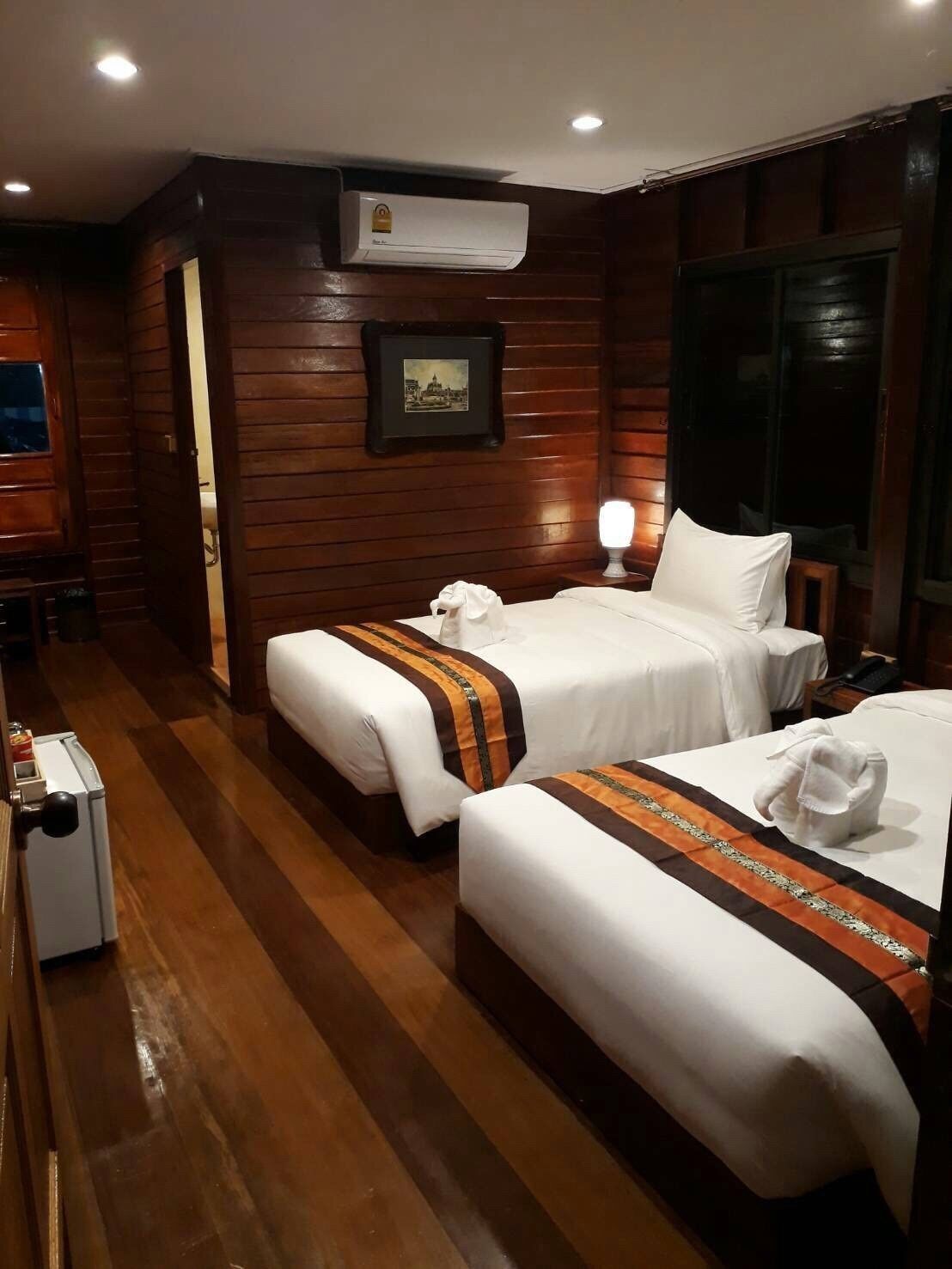 A hotel room with two beds, wooden paneling, an air conditioning unit, a framed picture on the wall, and a bedside lamp.