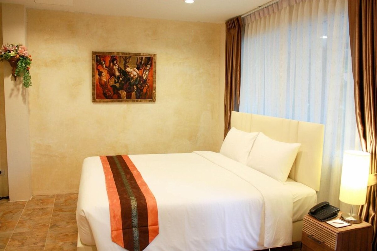 A hotel room with a large bed, a nightstand with a lamp, a painting on the wall, and a window with curtains.