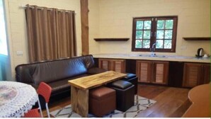 A compact living space with a sofa, a small wooden table, a kitchen area with a window, and a red chair. - Treasure Inn Khaoyai (Pak Chong)