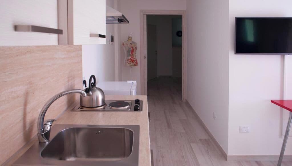 A modern kitchen with a stainless steel sink, a kettle, and a flat-screen TV mounted on the wall.
