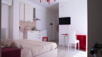 A bedroom with a bed, a desk, and a chair. There is a kitchenette with a stove and a sink.