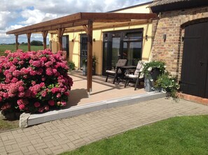 A covered patio area with outdoor seating, a brick wall, and a large flowering bush. - Quiet apartment with terrace and water views (Garz/Rügen)