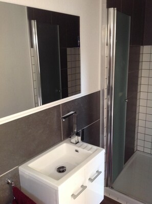 A bathroom with a white sink, a mirror, a glass shower enclosure, and dark tiled walls. - Quiet apartment with terrace and water views (Garz/Rügen)