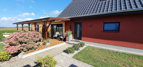 A red house with a tiled roof, a covered patio with outdoor furniture, and a garden with pink flowers. - Quiet apartment with terrace and water views (Garz/Rügen)