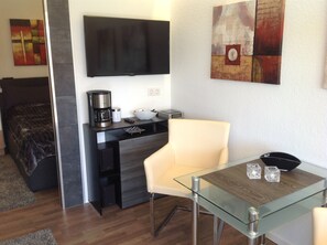 A modern hotel room with a bed, a desk with a coffee maker, a TV, and a glass table with decorative items.