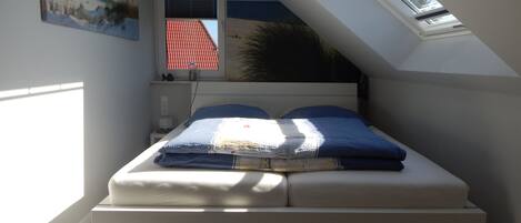 Ein Schlafzimmer mit schrägem Dach, einem Bett mit blau-weißer Decke, einem Fenster mit Blick auf ein rotes Dach und einem Bild an der Wand.