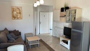 A modern living room with a sofa, a coffee table, and a TV stand. There is a kitchen area with a refrigerator, a microwave, and a cabinet.