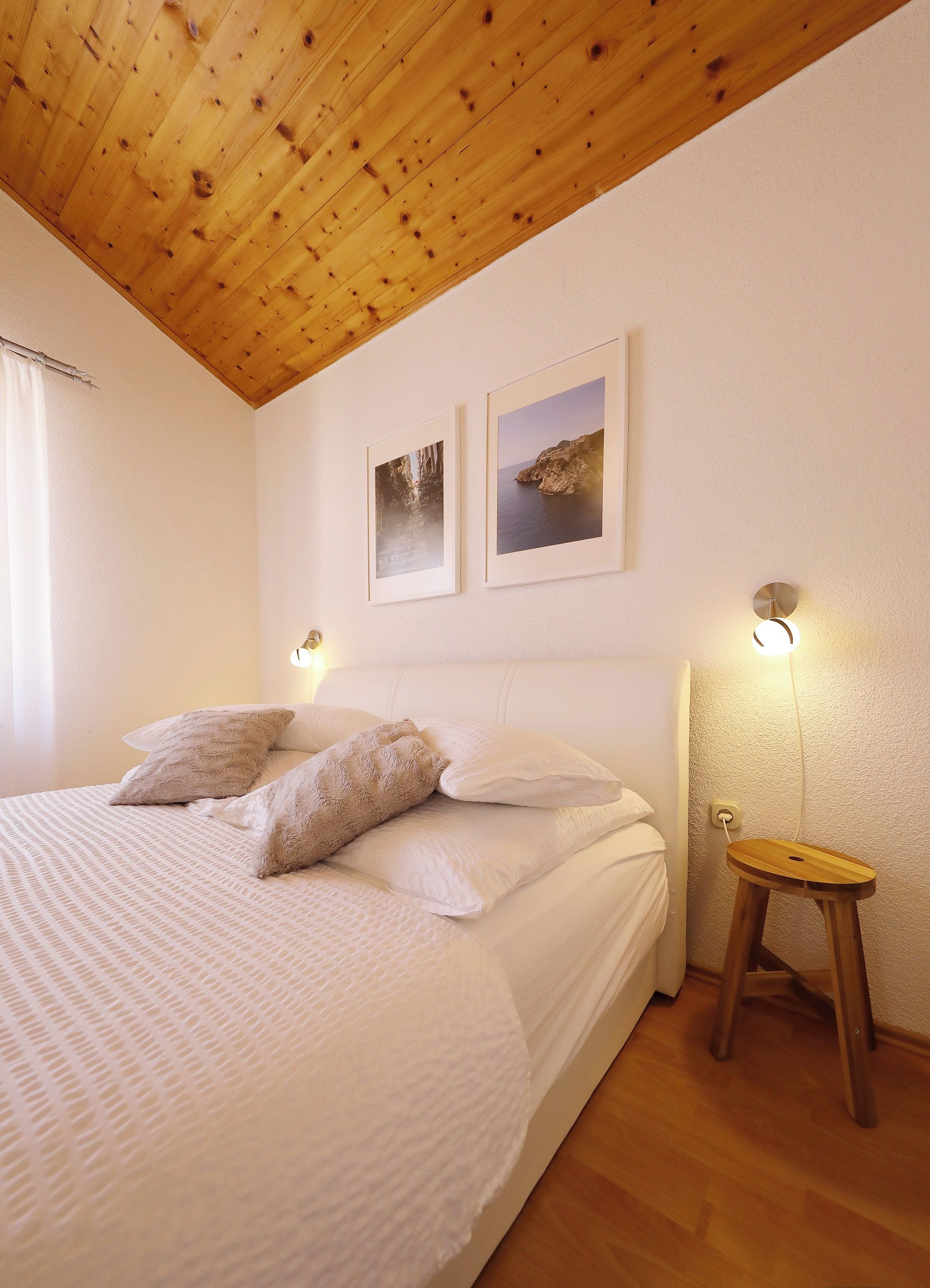A bedroom with a wooden ceiling, a bed with white bedding, two wall-mounted lights, and two framed pictures on the wall.