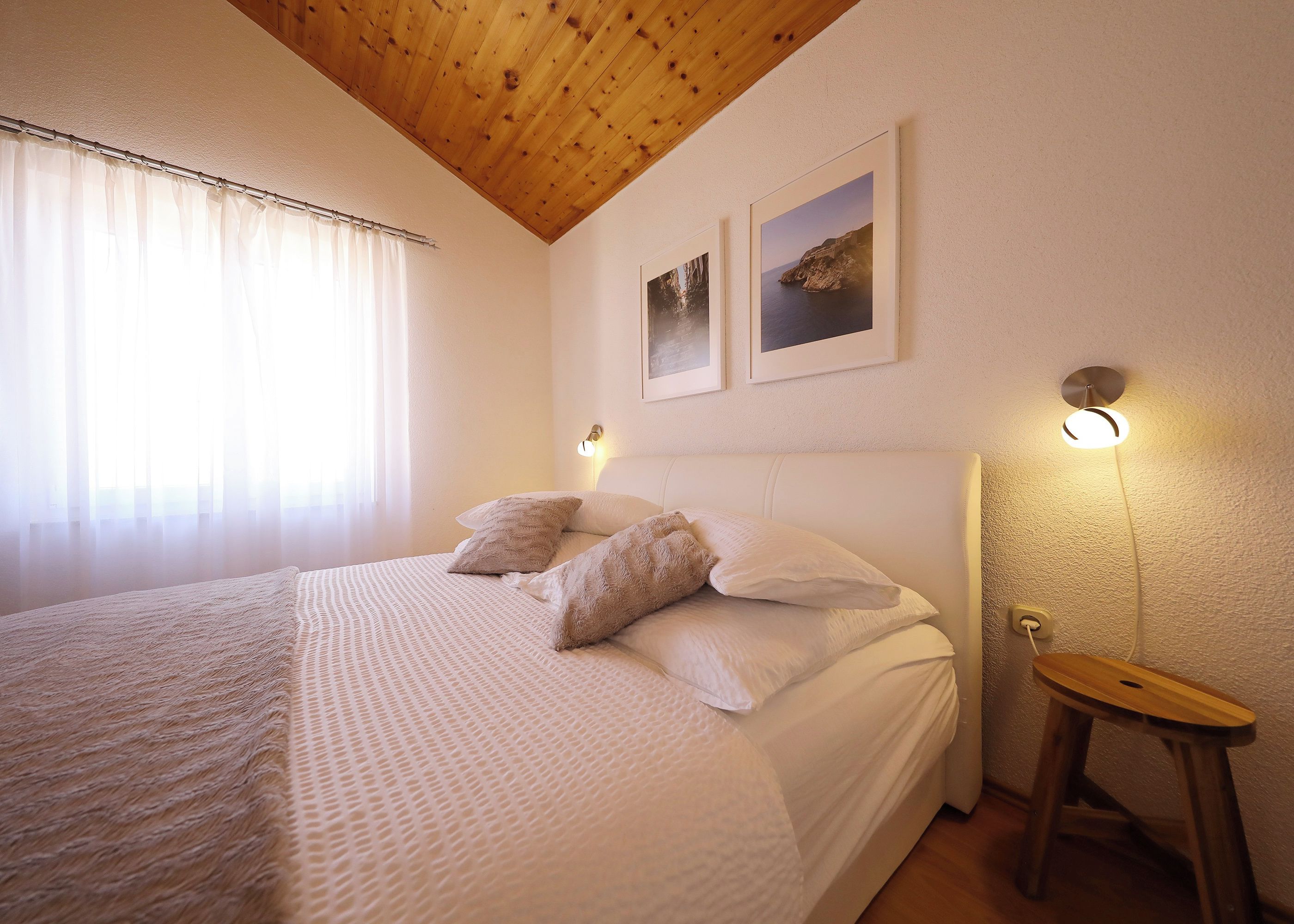 A bedroom with a large bed, wooden ceiling, two wall-mounted lights, and a small wooden stool.