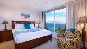 A bedroom with a large bed, a ceiling fan, a view of the ocean, and a chair with a floral pattern.
