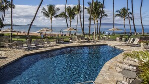 A pool area with lounge chairs and umbrellas, surrounded by palm trees, overlooking a beach and ocean.