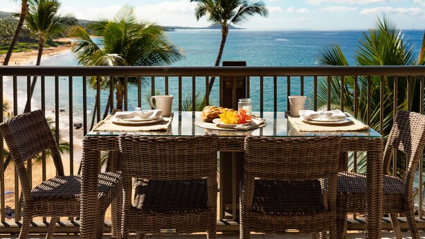 A balcony with wicker chairs and a table set for breakfast, overlooking a beach and ocean.