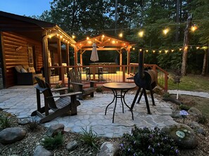 Terraza de madera con mobiliario exterior, una parrilla y luces colgantes.