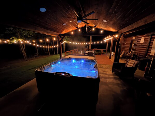 Un jacuzzi con luces azules, rodeado por una terraza de madera, luces colgantes y un área de estar cubierta.