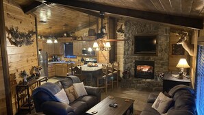 A cozy log cabin interior with a stone fireplace, wooden furniture, and a kitchen area.