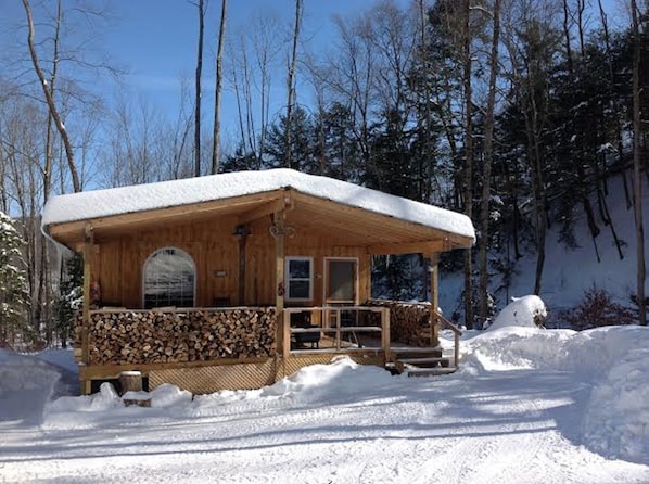 Eine Holzhütte mit schneebedecktem Dach, einer Veranda mit gestapeltem Holz und freiem Blick auf den umliegenden Wald.